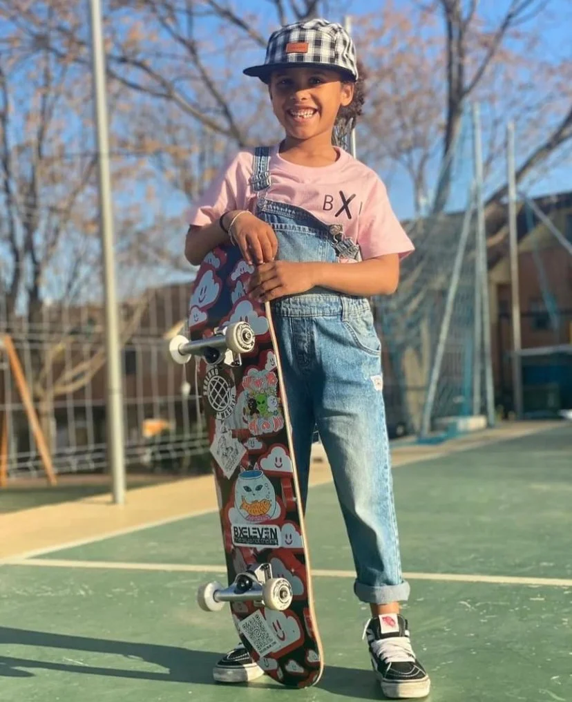 A young girl with curly hair wearing a checkered print cap, pink t-shirt, and denim overalls, smiling and standing on a skateboard outdoors on a sunny day